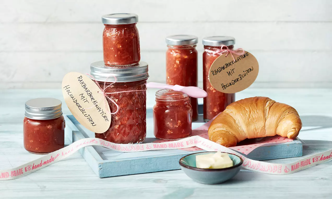 Rhabarberkonfitüre in gläsernen Einmachgläsern mit silbernen Deckeln und handgeschriebenen Etiketten auf hellem Holztablett mit Croissant