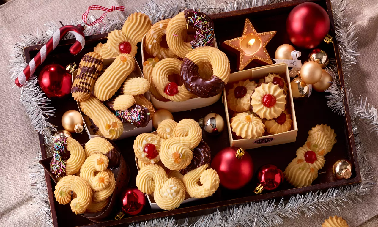 Spritzgebäck in heller, leicht gebräunter Farbe mit geschwungenen Mustern, teils mit Schokolade oder Kirschen verziert, auf weihnachtlich dekorierter Platte