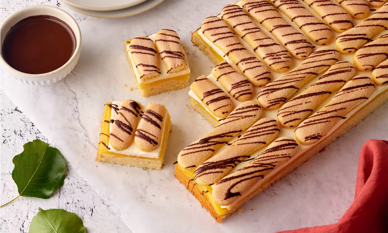 Lambadaschnitten mit gelber Creme und heller Biskuitschicht, dekoriert mit dunklen Schokostreifen, arrangiert auf weißem Untergrund
