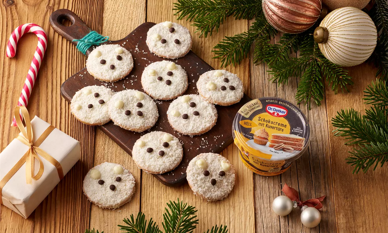 Runde Kekse mit weißer Kokosschicht, dunklen Schokopunkten als Augen und Nase, angerichtet auf Holzbrett mit Tannenzweigen und Weihnachtsdeko