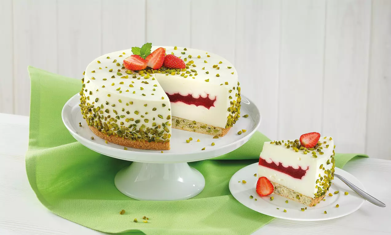Runde Joghurt-Torte mit weißer Creme, roter Erdbeerfüllung und gehackten Pistazien, auf Tortenplatte und grünem Tuch angerichtet