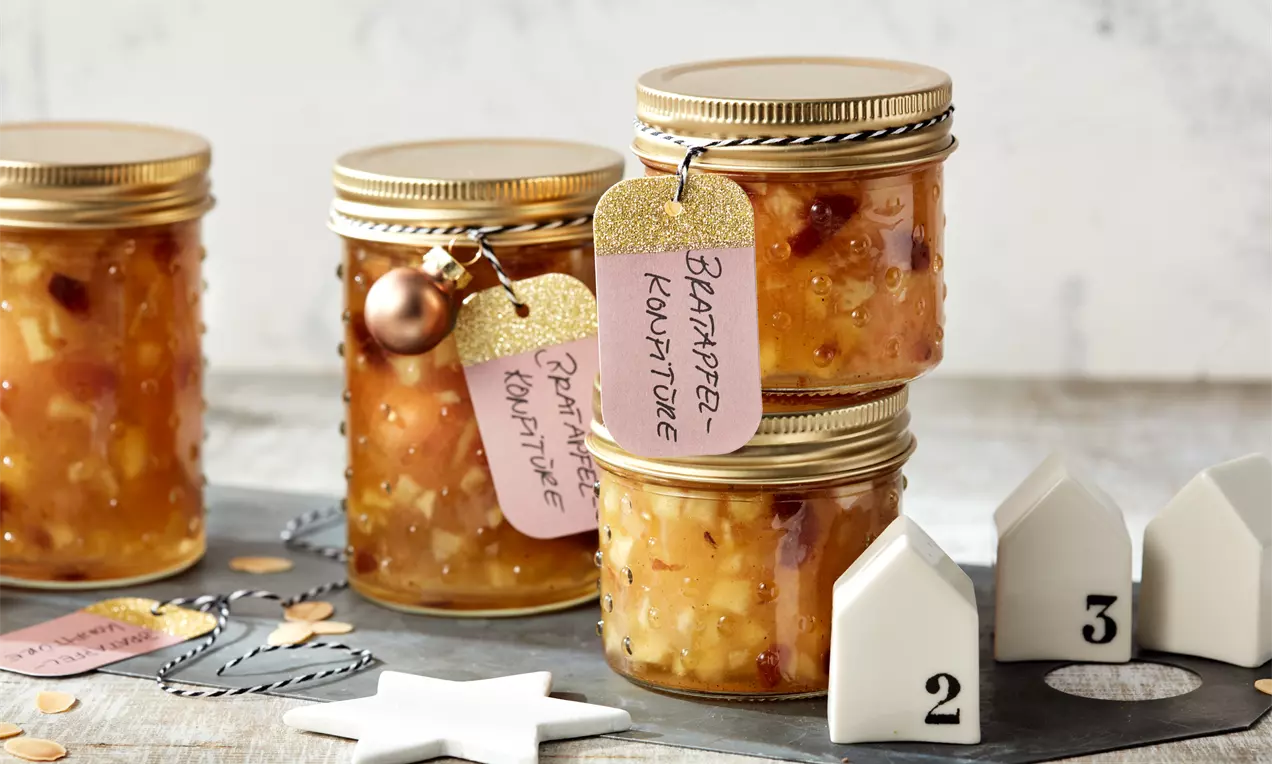 Vier goldbraune Gläser Bratapfel-Konfitüre mit goldenen Deckeln und rosa Anhängern auf dunklem Tisch mit weißen Dekoelementen