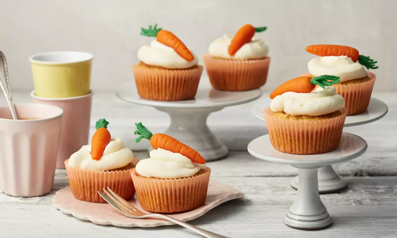 Rübli-Cupcakes mit beiger Frischkäsecreme und orangefarbenen Marzipanmöhren auf weißen Tortenständern und rosa Tellern arrangiert