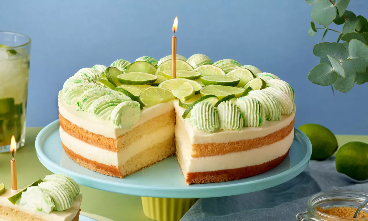 Mojito-Torte mit heller Creme und goldbraunen Biskuitböden, dekoriert mit grünen Sahnetupfen und frischen Limettenscheiben auf hellblauer Tortenplatte