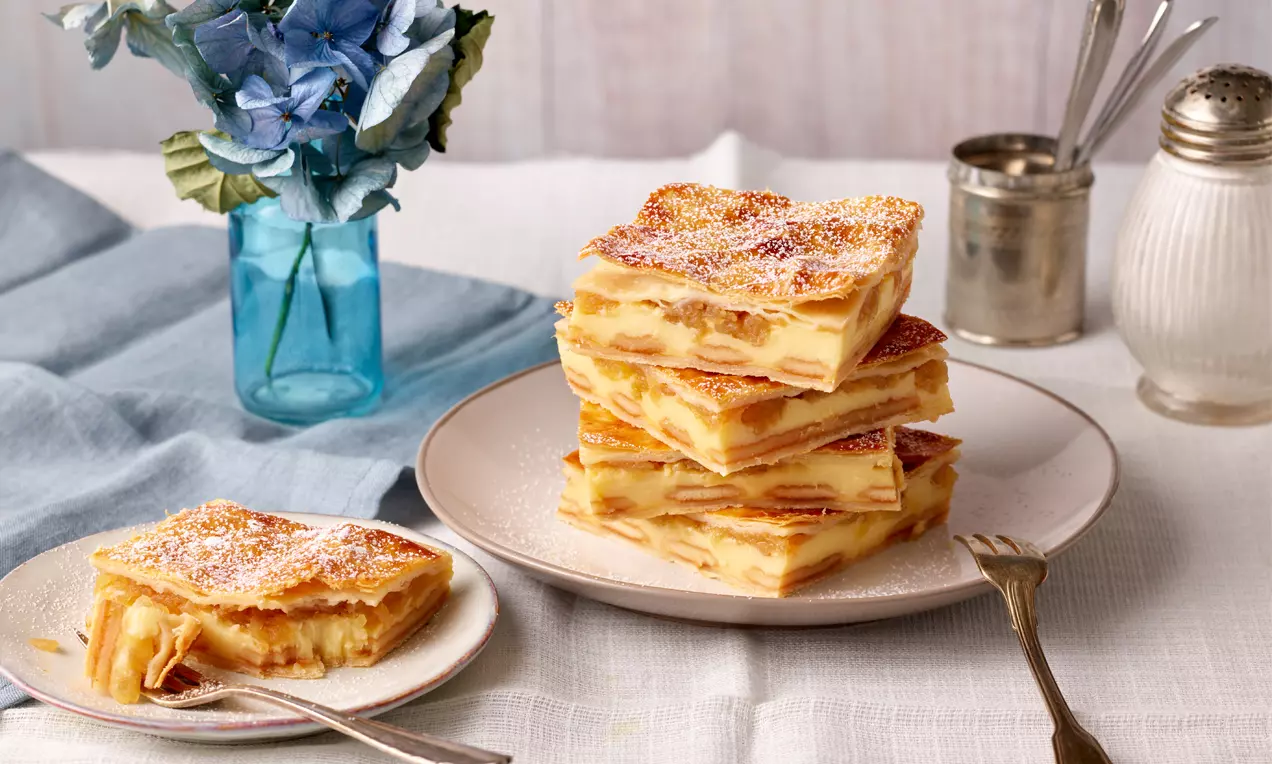 Apfel-Cremeschnitten mit goldbrauner Blätterteigkruste und cremiger Füllung als Stapel auf einem Teller, daneben eine einzelne Portion auf kleiner Platte