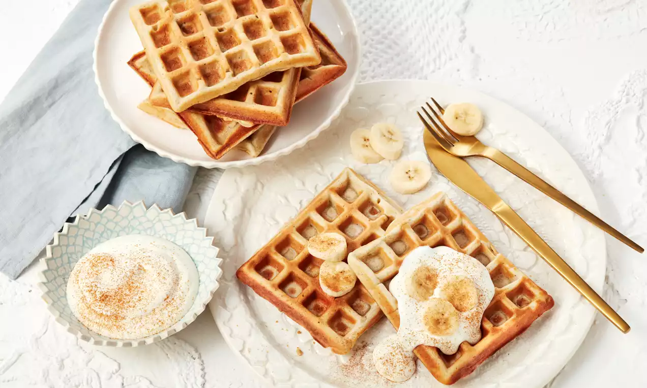 Quadratische, goldbraune Bananenwaffeln mit weicher Textur auf weißem Teller, garniert mit Bananenscheiben und Sahne, daneben goldenes Besteck