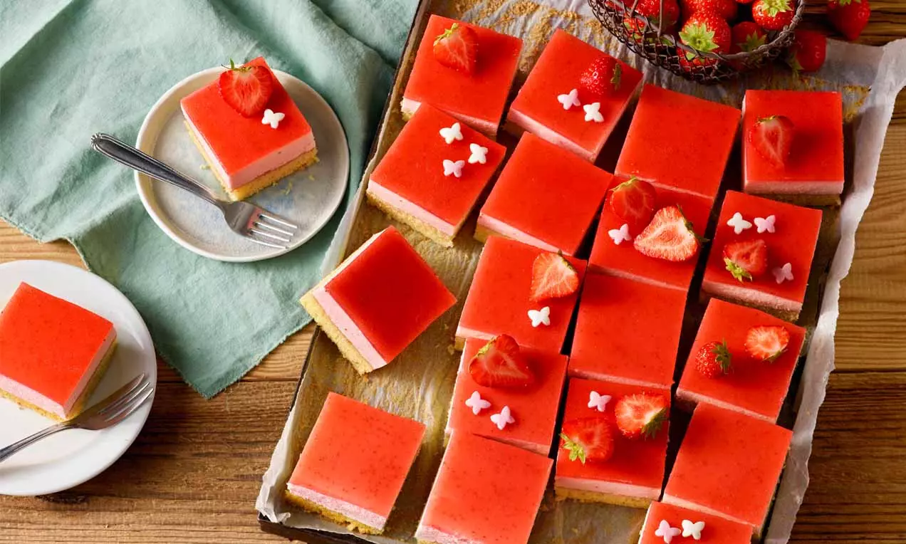 Kleine quadratische Stücke Erdbeer-Blechkuchen mit roter glänzender Gelee-Schicht und Deko aus weißen Zuckerblumen und geschnittenen Erdbeeren