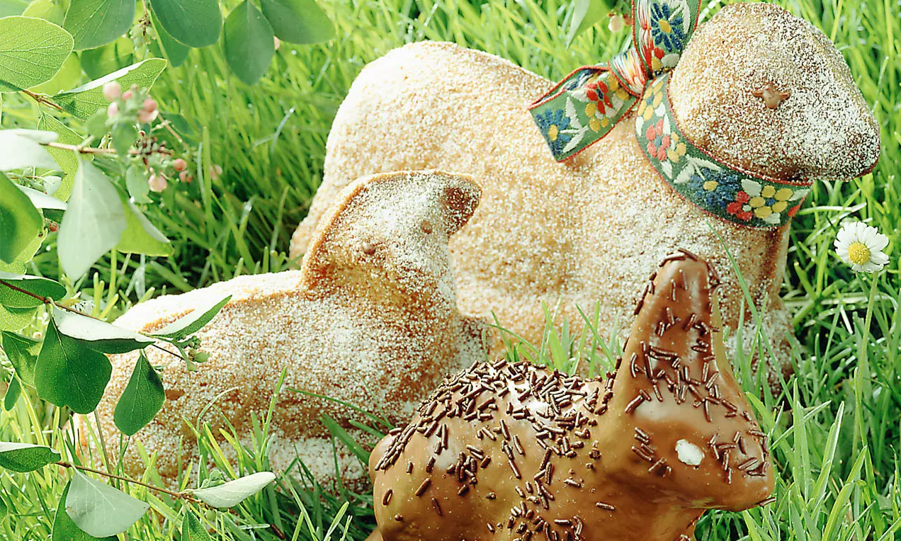 Gebäck-Osterlämmer mit goldbrauner Kruste und Puderzucker, verziert mit Schleife, vor Osterhase mit Schokoglasur und Streuseln im Gras