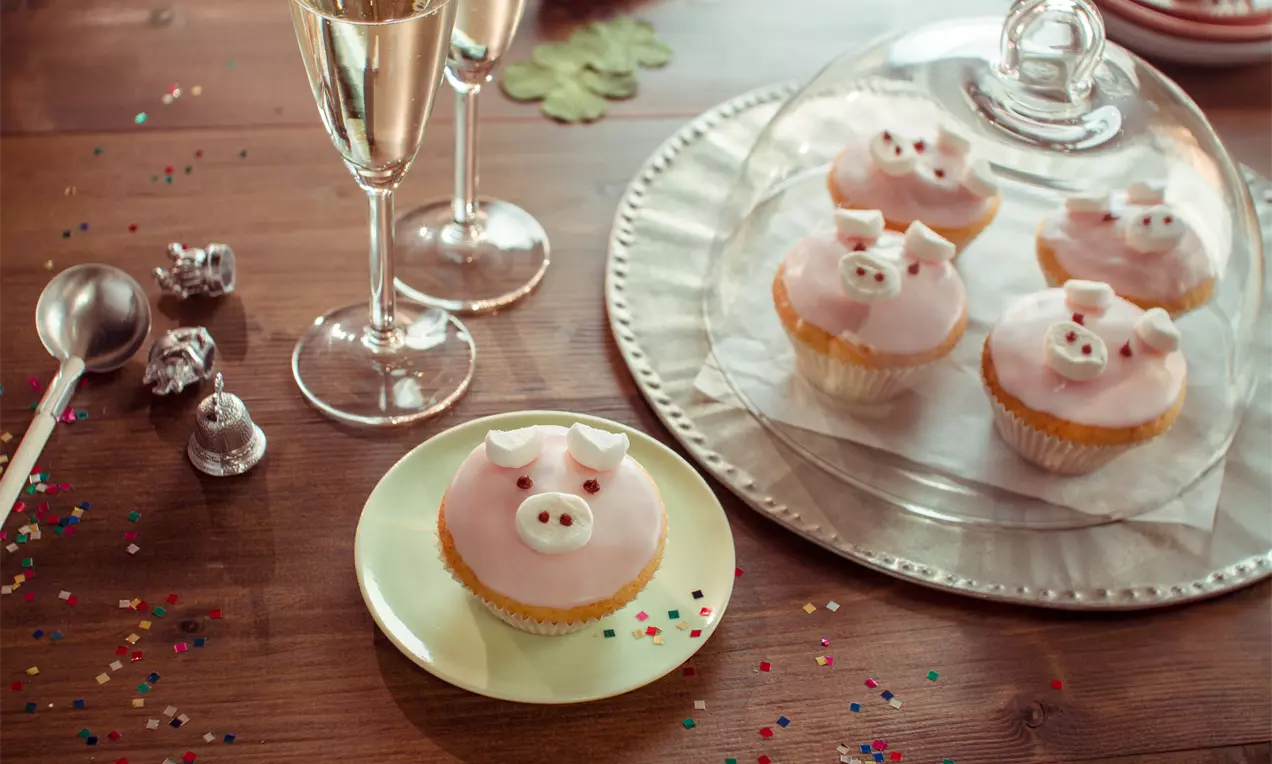 Rosa verzierte Cupcakes mit Schweinegesichtern, kleine Ohren und Nasen aus Fondant, arrangiert unter einer Glashaube auf weißem Teller