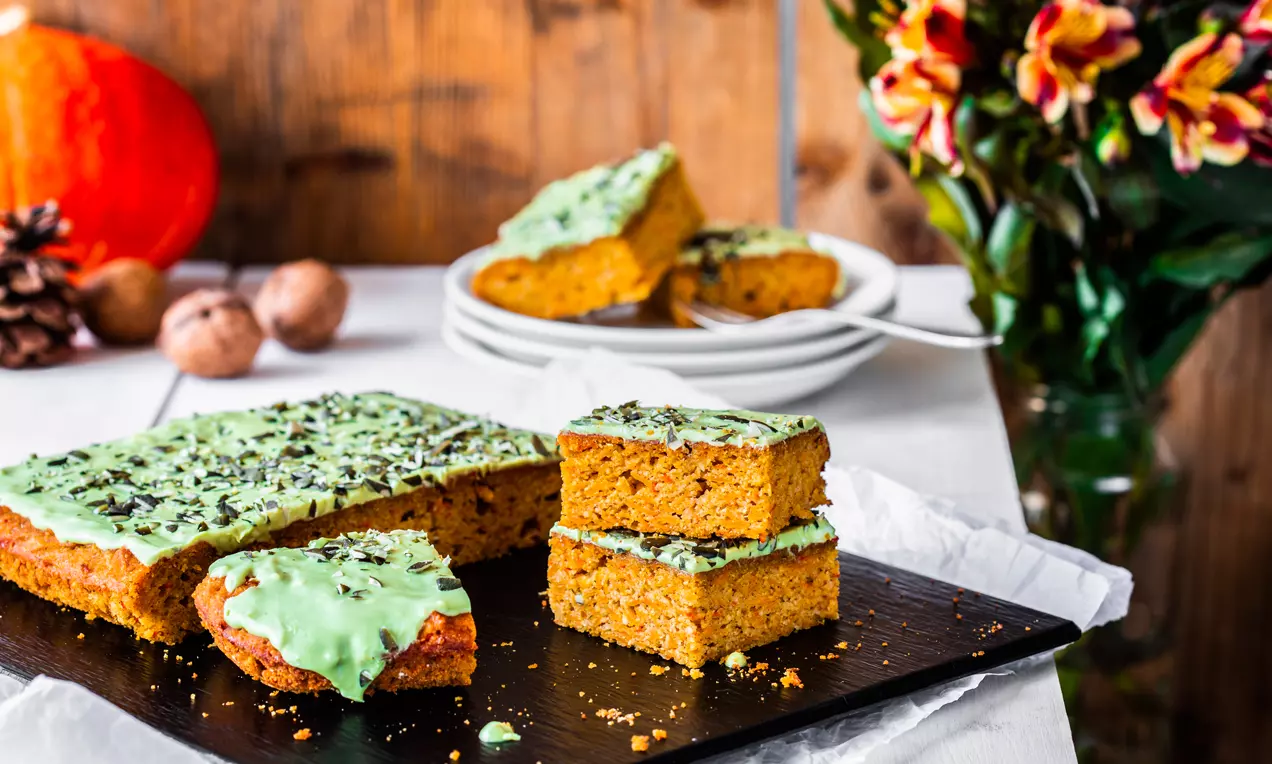 Glutenfreie Kürbis-Schnitten mit orangefarbener, lockerer Textur, hellgrüner Glasur und dunklen Schokosplittern auf einem schwarzen Holzbrett
