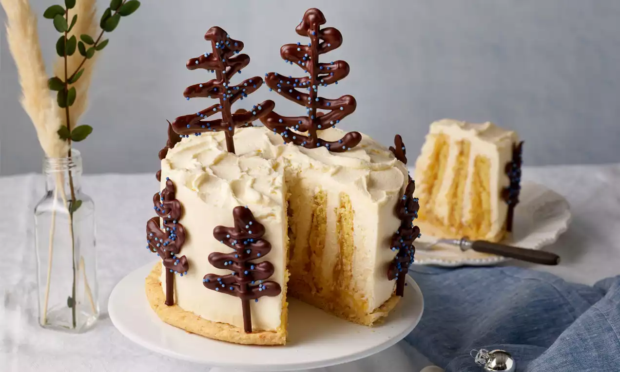 Weiße Weihnachtstorte mit heller Creme, Schokoladentannen und Sicht auf goldgelbe Schichten, auf einer Tortenplatte serviert
