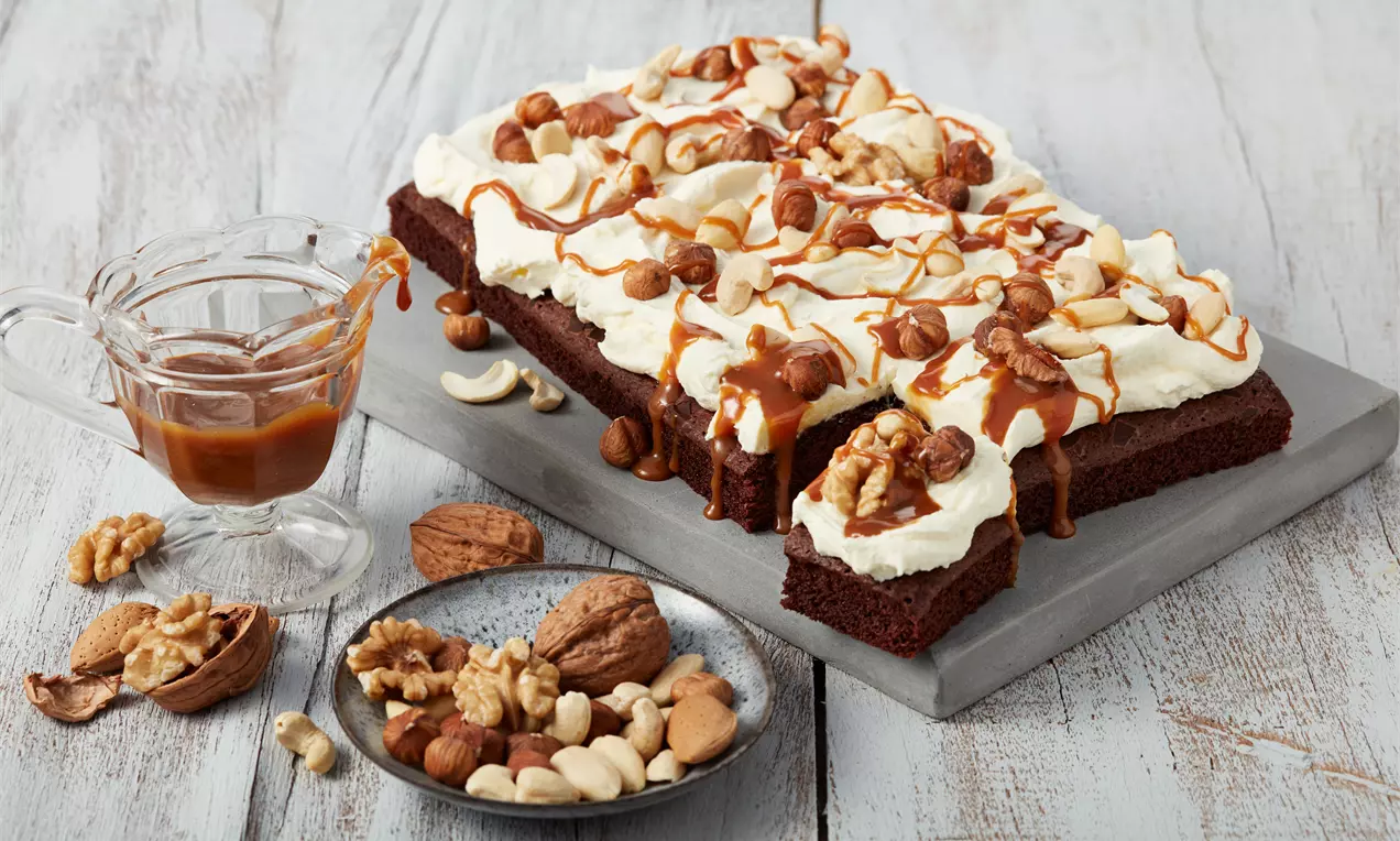 Saftiger, dunkelbrauner Brownie mit luftigem Frischkäse-Topping, Karamell-Drizzle und knackigen Nüssen auf grauer Platte mit Beilagen