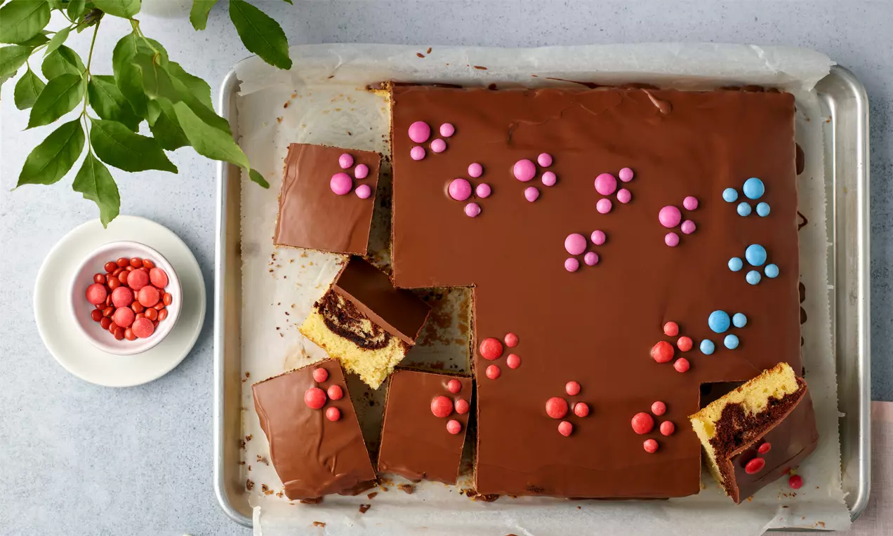 Flacher Marmorkuchen vom Blech mit glänzender Schokoglasur, bestreut mit bunten Schokolinsen und angeschnitten auf einem Backpapier
