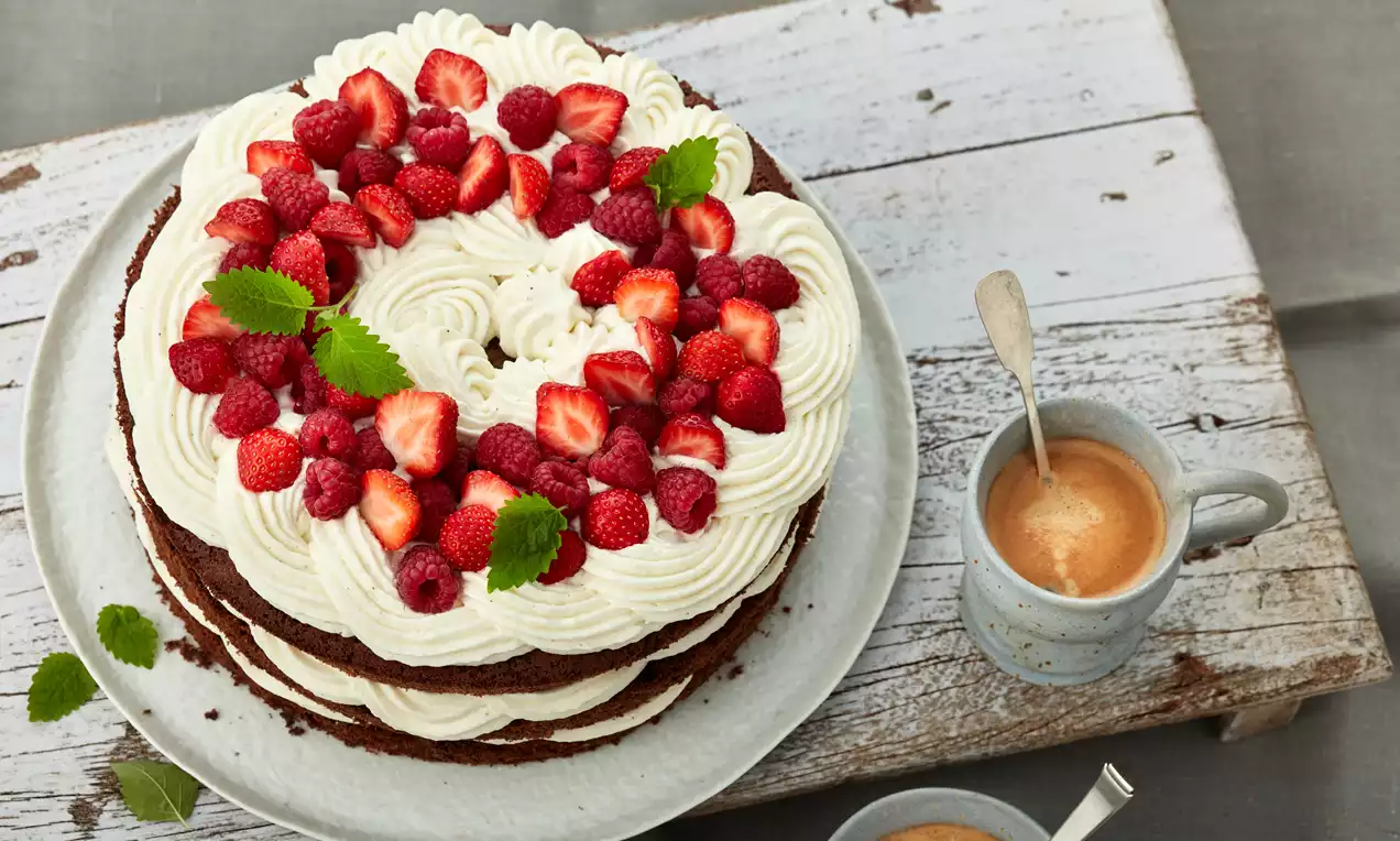 Erdbeer-Himbeer-Torte mit weißer Sahnecreme in geschwungenen Mustern, rotem Beerenbelag und grünen Blättern auf grauem Teller