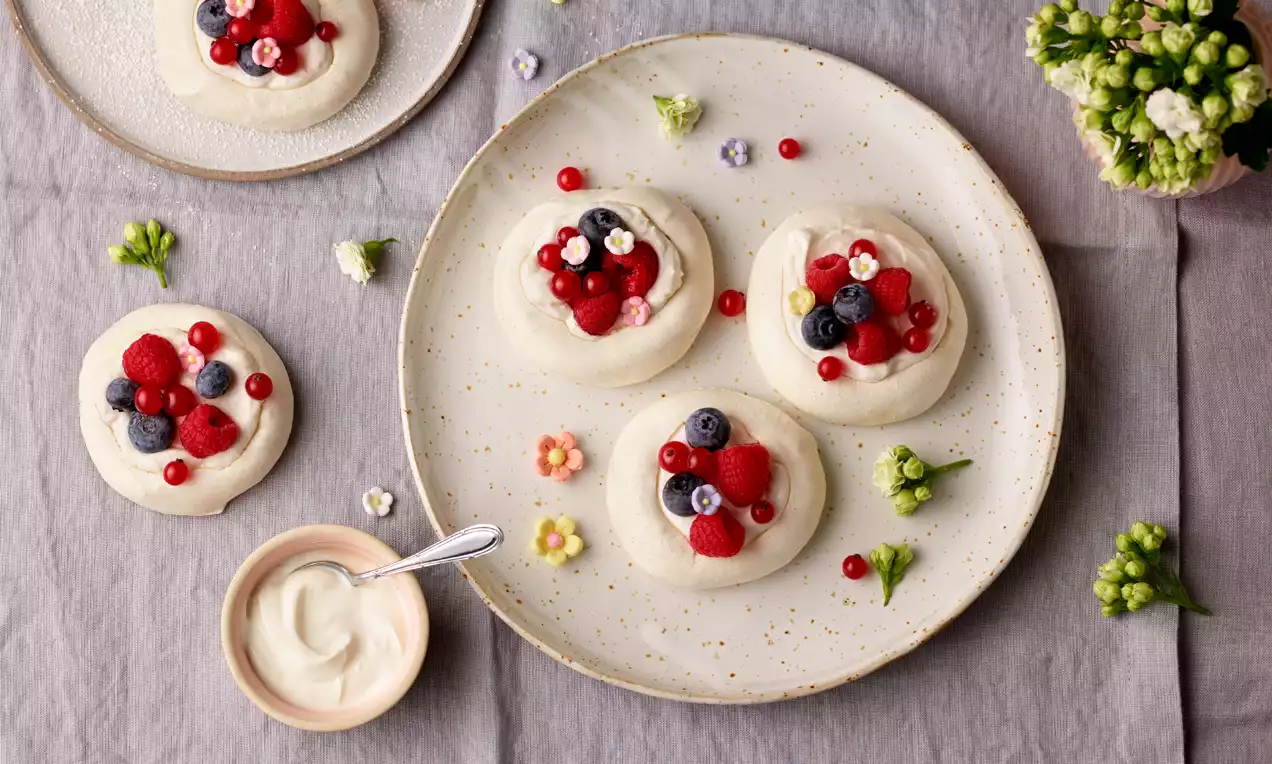 Mini-Pavlova mit lockerer, weißer Cremefüllung, saftig roten Himbeeren, tiefblauen Blaubeeren und weißen Blüten auf einem hellen Teller drapiert