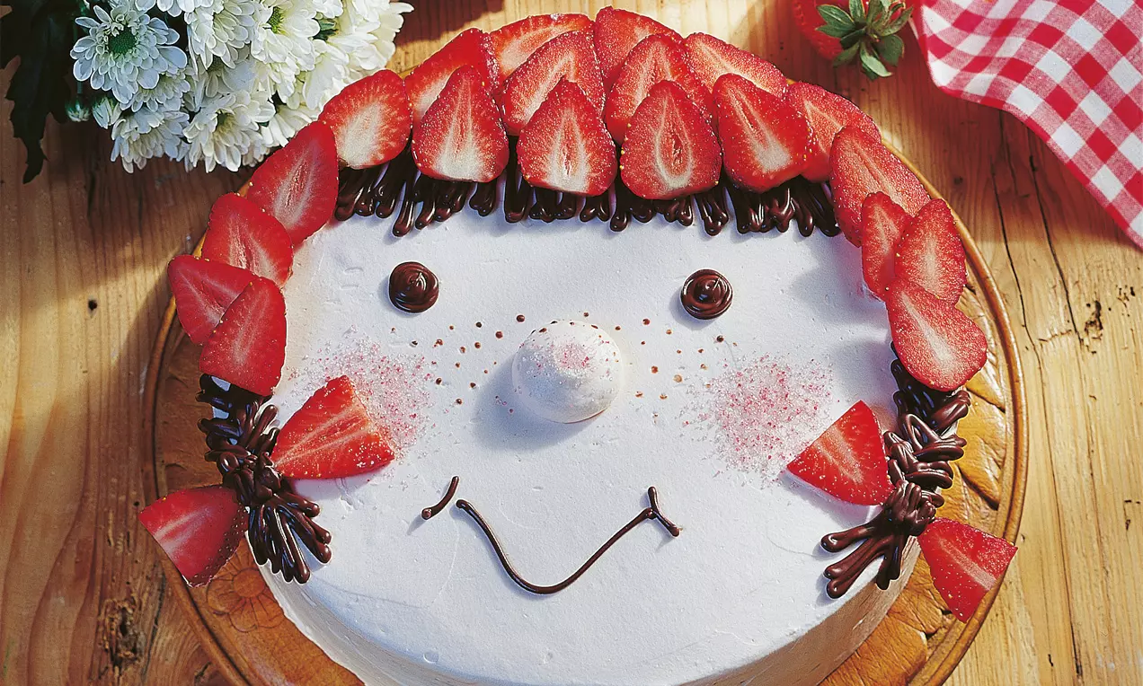 Rotkäppchen-Torte mit weißer Creme, Erdbeeren als Haare, Schokoladen-Dekor als Gesichtszüge auf einem Holzteller drapiert