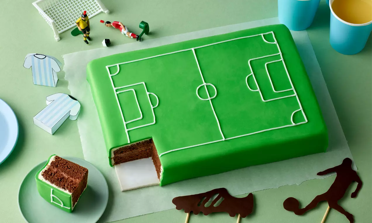 Rechteckige Torte mit glatter grüner Fondantdecke und weißem Spielfeldmuster, ein aufgeschnittenes Stück zeigt Schokoladenbiskuit