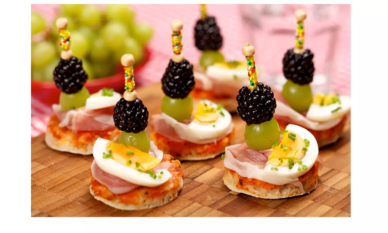 Kleine runde Brotstückchen mit Prosciutto, Ei, Schnittlauch und dekorativen Spießen aus Brombeeren und grünen Weintrauben auf Holzbrett