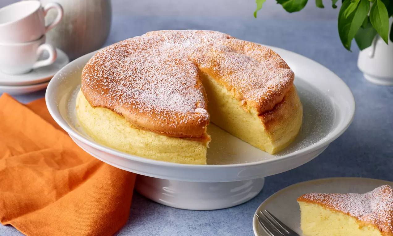 Lockerer japanischer Käsekuchen mit goldener, leicht gebräunter Oberfläche und Puderzucker bestäubt auf weißer Kuchenplatte mit angeschnittenem Stück