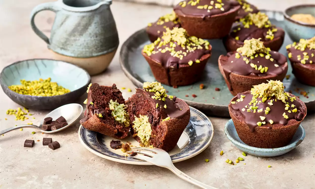 Schokoladenmuffins mit glänzender Glasur und gehackten Pistazien, auf Tellern arrangiert neben einer Kanne und kleinen Schälchen