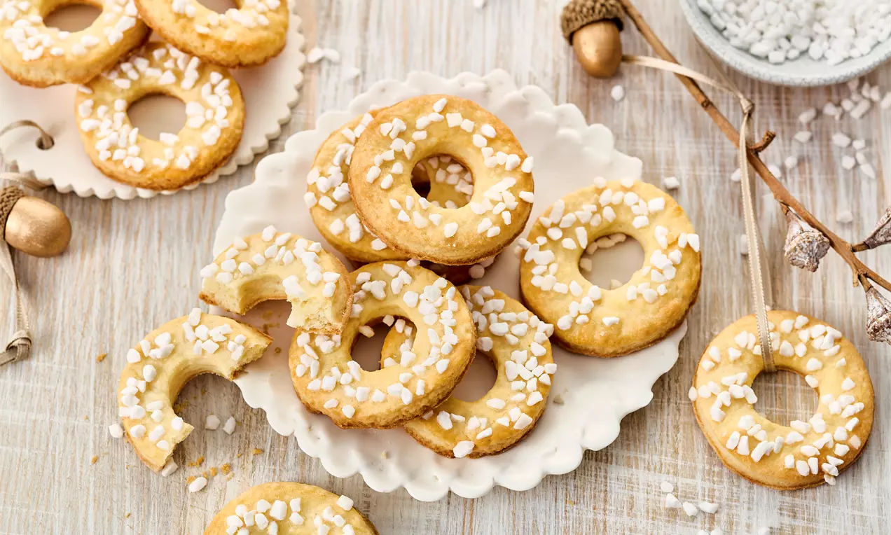 Goldbraune, ringförmige Saure Sahne-Plätzchen mit weißem Hagelzucker auf einem zarten Teller, umgeben von herbstlicher Dekoration
