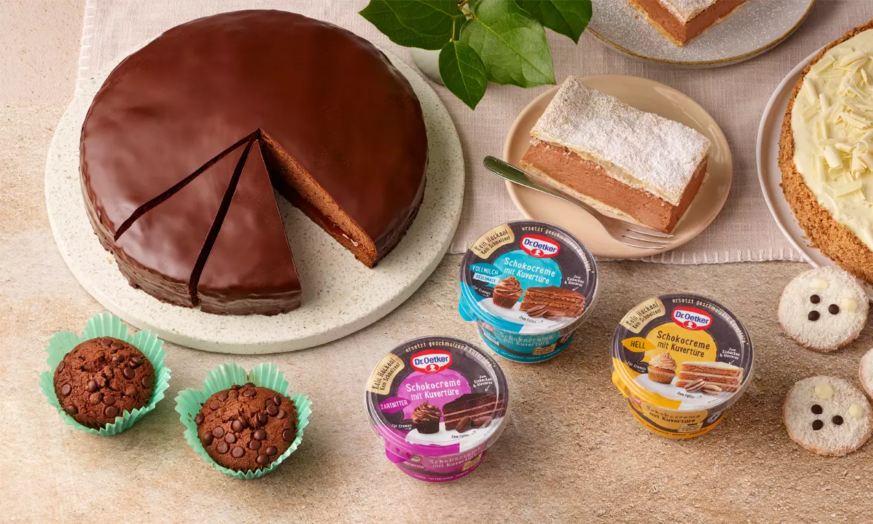 Ein runder Schokoladenkuchen mit glänzendem Überzug und angeschnittenem Stück auf einem hellen Teller, umgeben von Desserts und Muffins