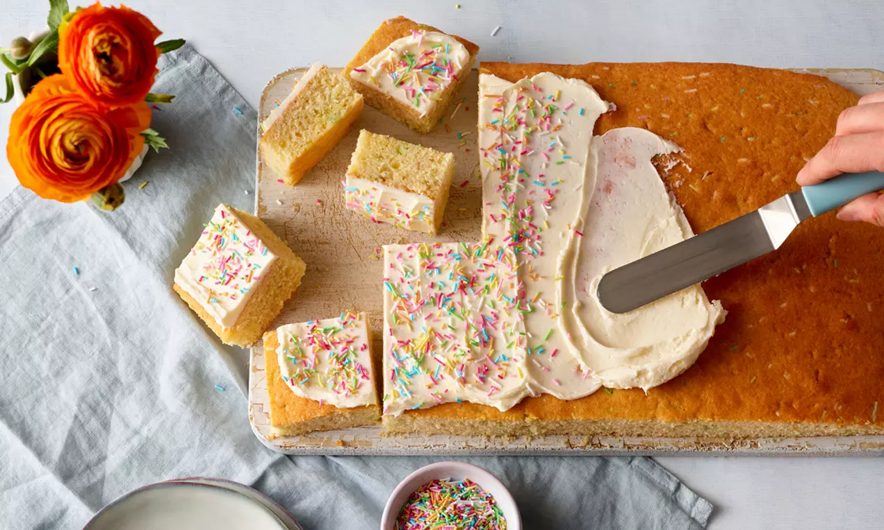 Rechteckiger heller Kuchen mit weißer Creme und bunten Streuseln, teils in Stücke geschnitten, auf Holzbrett mit gelben Ranunkeln im Hintergrund