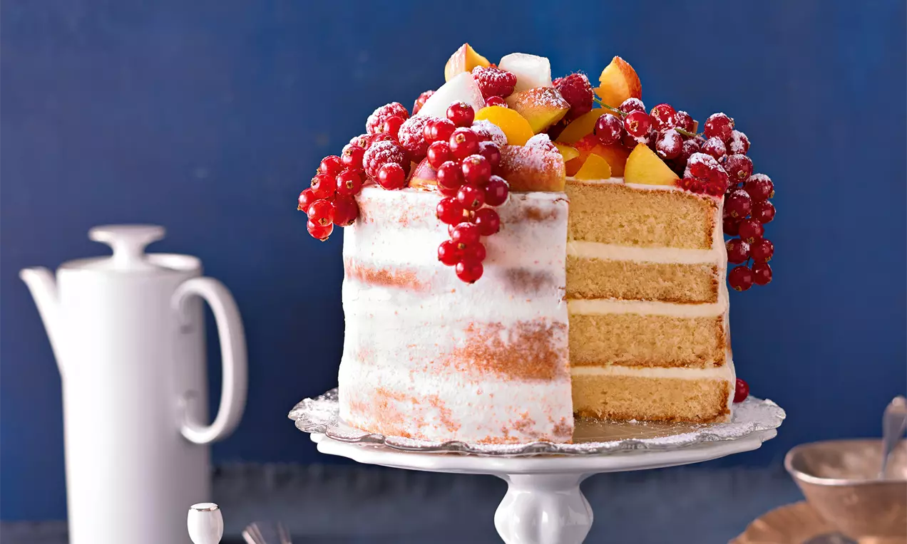 Hohe Schichttorte mit heller Creme und luftigem Bisquitteig, gekrönt von roten Johannisbeeren, Pfirsichstücken und Puderzucker, auf einer Tortenplatte.