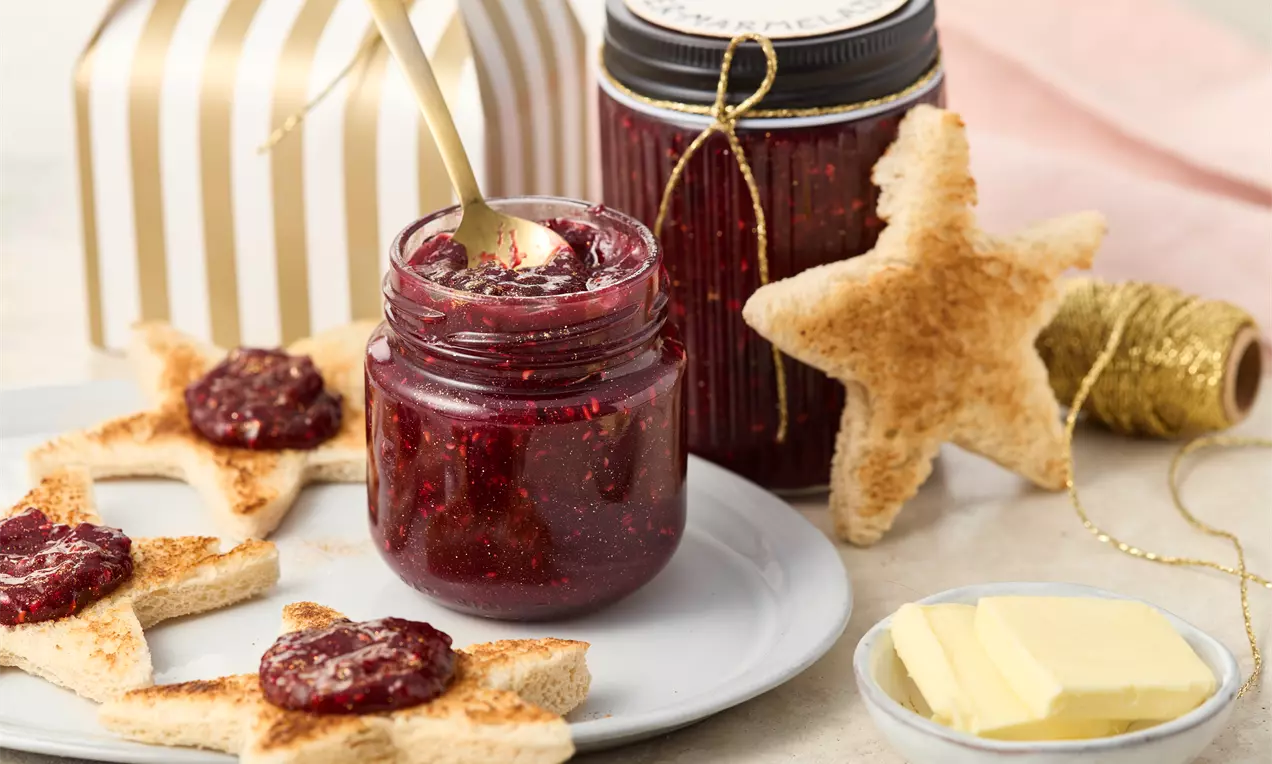 Rot-glänzende Marmelade in einem Glas mit goldfarbenem Löffel, umgeben von sternförmigen Toastscheiben und Butterstücken auf einem Teller
