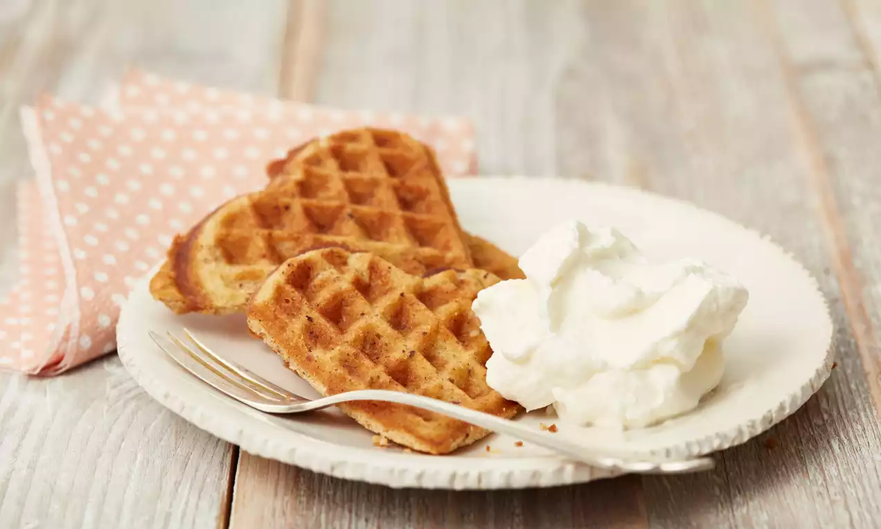 Herzförmige goldbraune Nuss-Waffeln mit feiner Textur auf cremeweißem Teller, daneben lockerer Sahneklecks und Besteck auf Holz-Tisch