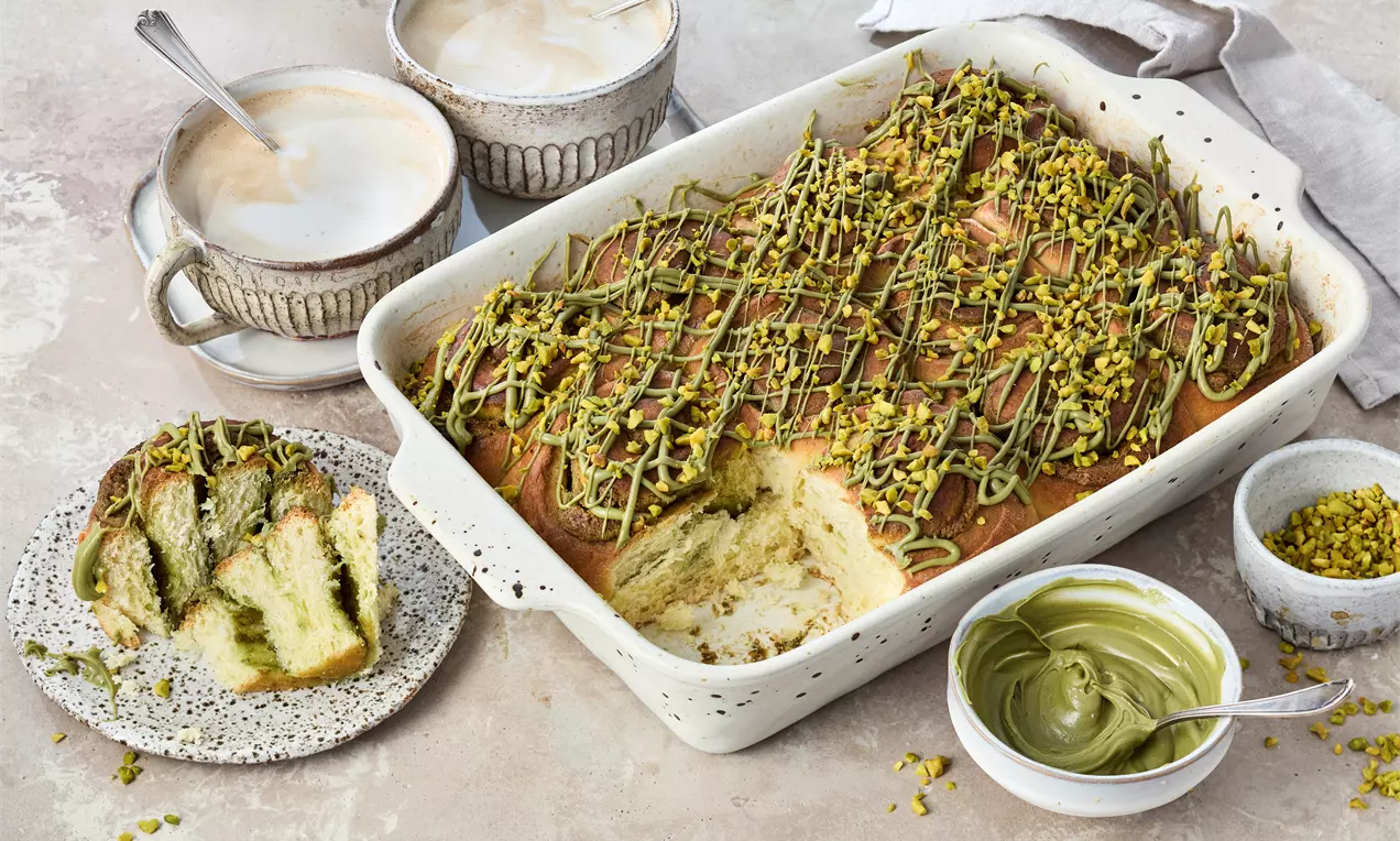 Goldbraune Pistazienschnecken in einer weißen Auflaufform, mit grünem Topping und gehackten Pistazien bestreut, daneben Kaffee und Pistaziencreme