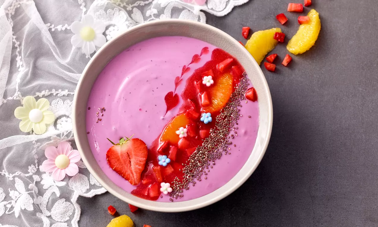 Cremiger pinker Joghurt in einer Schale, dekoriert mit Erdbeerwürfeln, Orangenspalten, Chiasamen und kleinen Zuckerdekoren