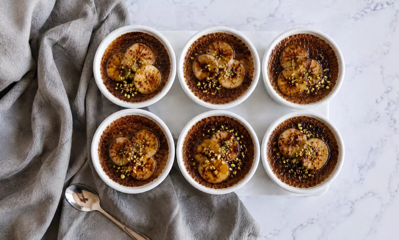 Schoko-Crème Brûlée mit dunkler, karamellisierter Kruste, belegt mit goldbraunen Honig-Bananenscheiben und gehackten Pistazien in weißen Förmchen, AI-labeled image