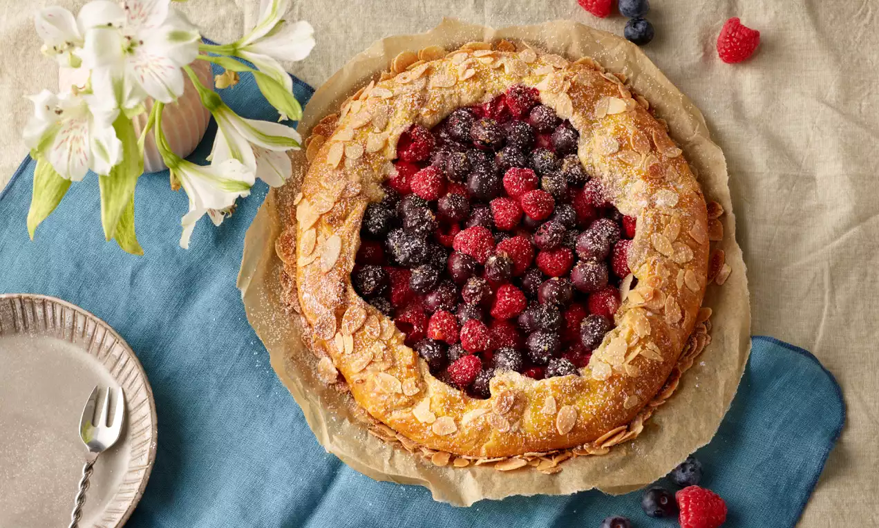 Goldbraune Crostata mit Buttermandelkruste, gefüllt mit glänzenden Himbeeren und Heidelbeeren, auf Backpapier und blauem Stoff serviert