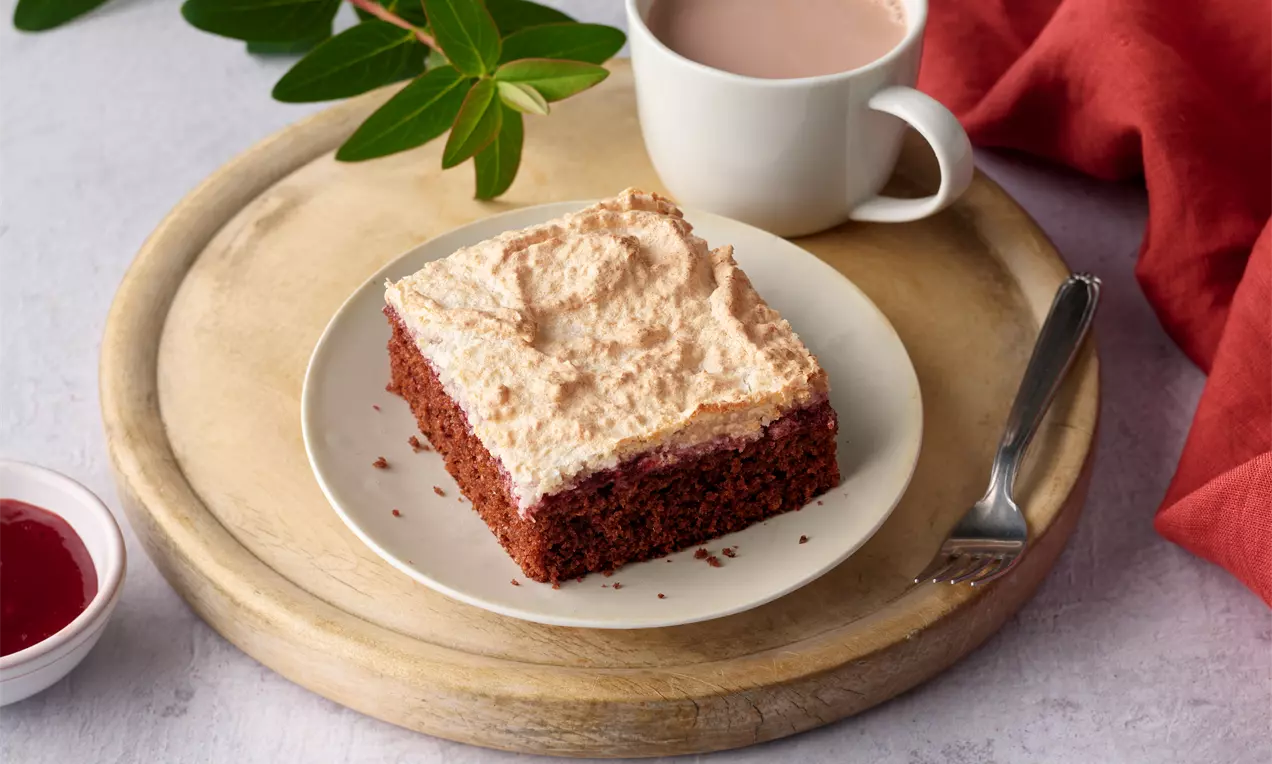 Saftiger Blechkuchen mit dunklem Teig und luftiger Kokoshaube, angerichtet auf einem weißen Teller mit Kaffee und rotem Tuch auf Holzbrett