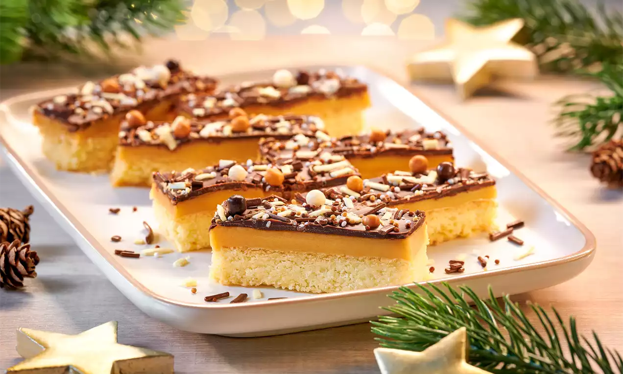 Rechteckiges Shortbread mit goldgelber Karamellschicht, glänzender Schokoglasur und bunten Streusel-Dekoren auf weißer Platte mit Tannenzweigen