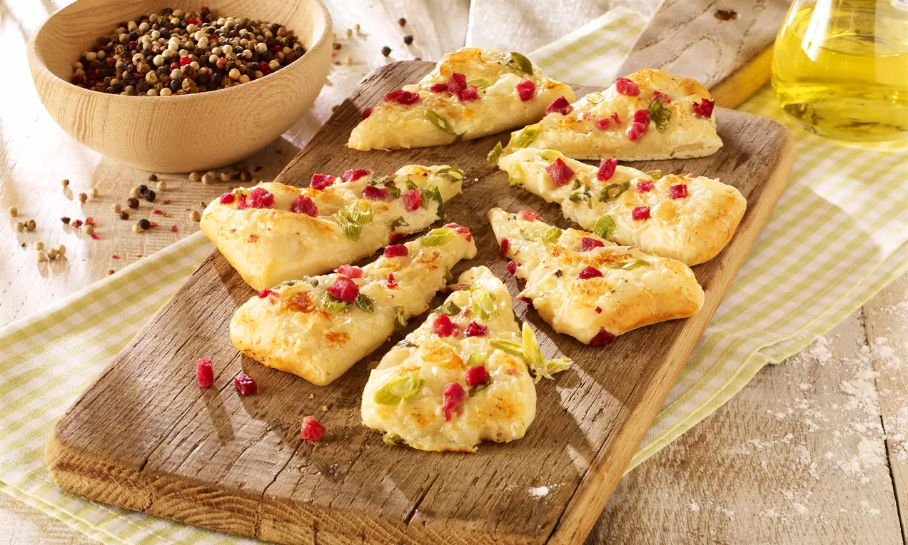 Goldbraune Schinkenecken mit geschmolzenem Käse und grünen Kräutern, bestreut mit roten Speckwürfeln auf rustikalem Holzbrett