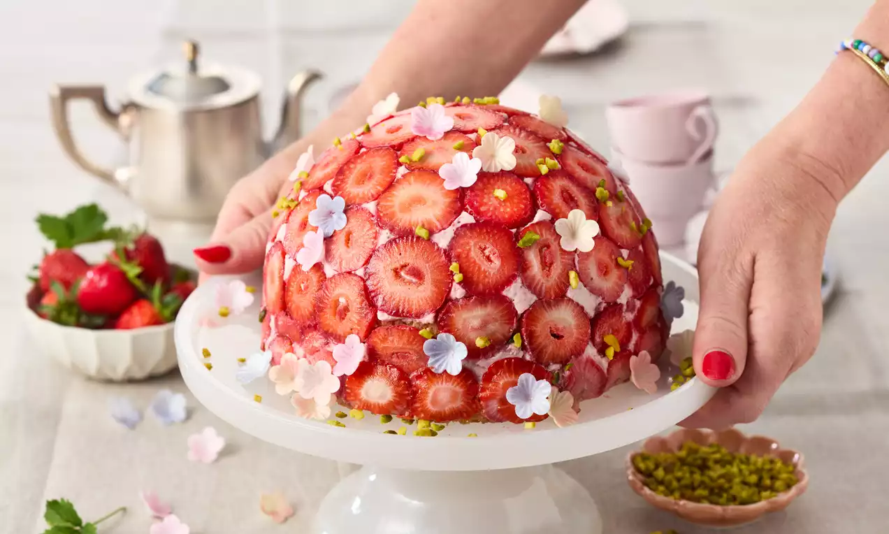 Kuppelförmige Erdbeertorte mit saftigen roten Erdbeerscheiben, bunten Zuckerblüten und gehackten Pistazien auf weißer Tortenplatte
