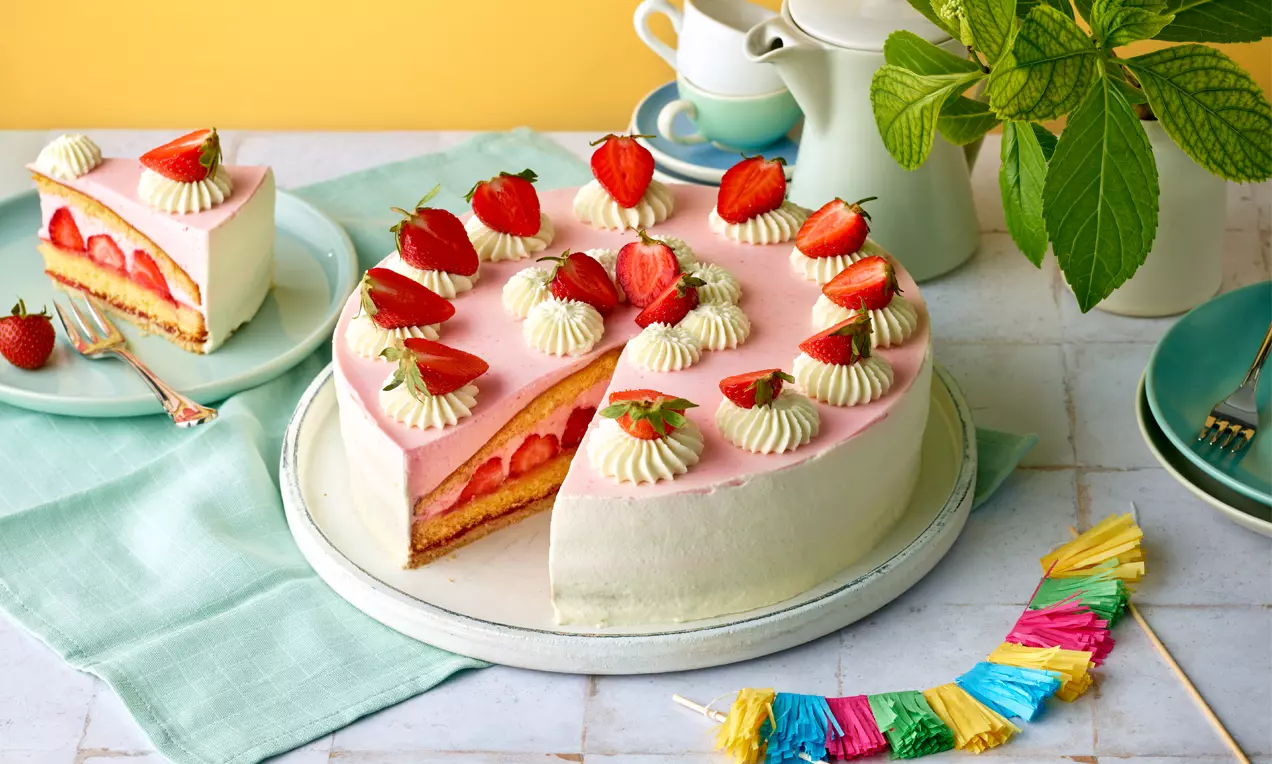 Erdbeer-Obers-Torte mit rosa Glasur, frischen Erdbeeren und Sahnetupfen auf weißem Teller, garniert mit bunten Papierdeko-Streifen