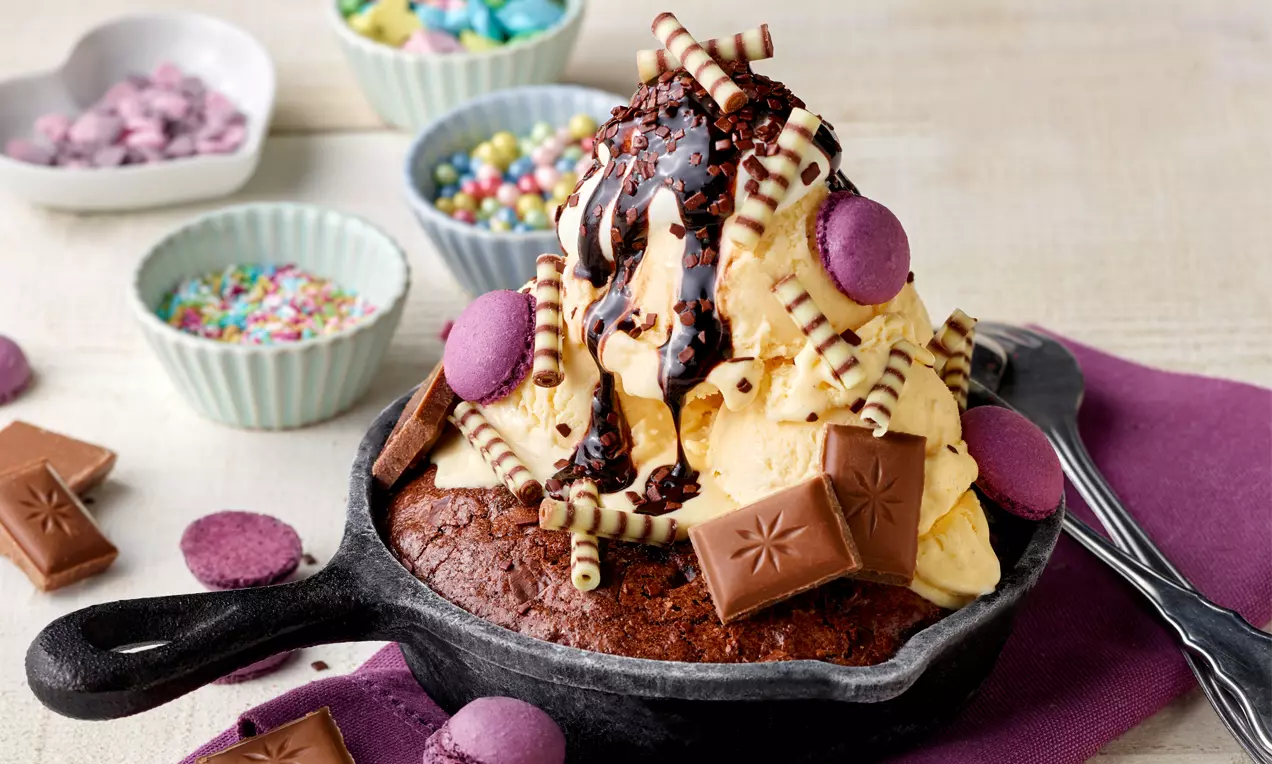 Ein warmes Brownie in einer schwarzen Eisenpfanne, belegt mit Vanilleeis, Schokoladensauce, lila Macarons, Schokolade und dekorativen Streuseln