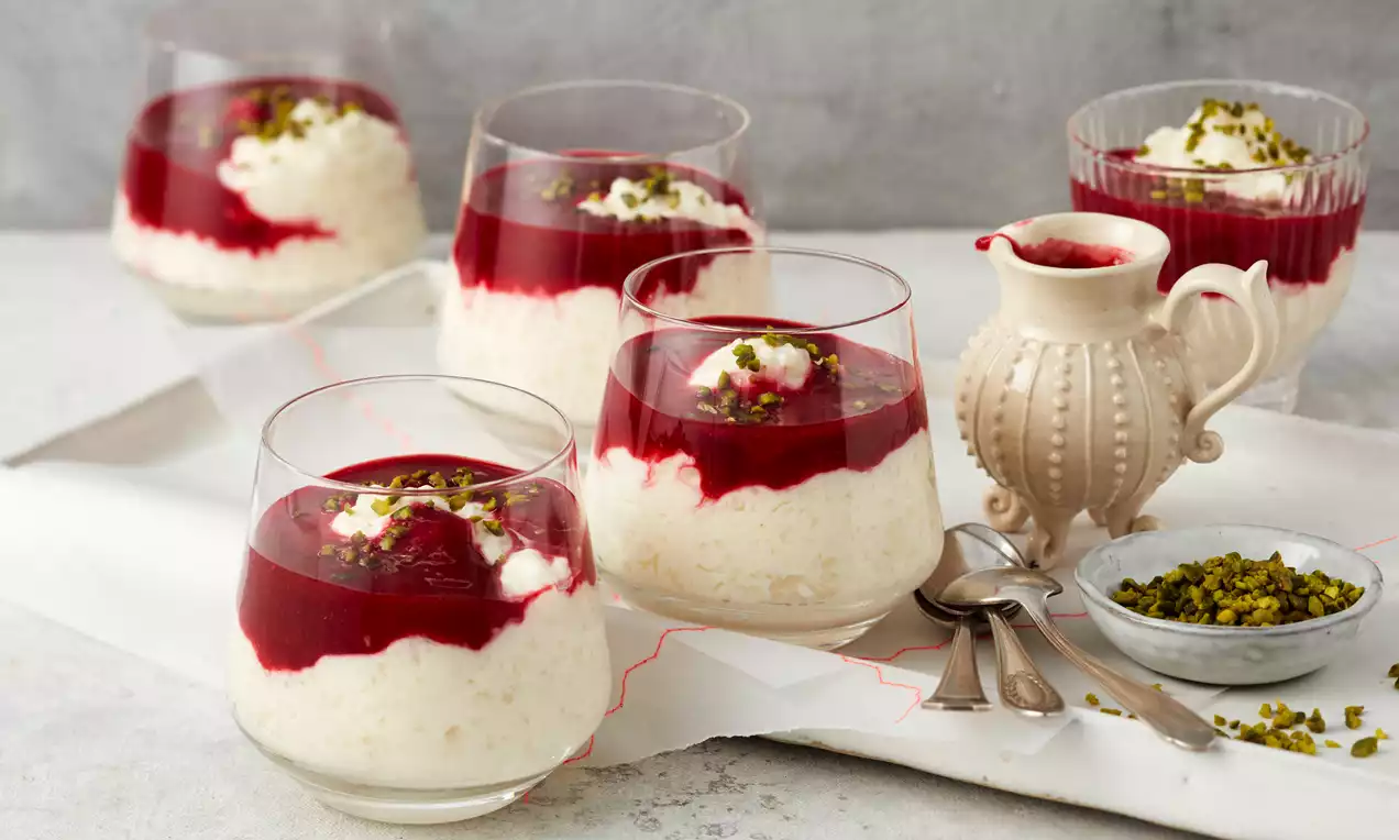 Gläser mit weißem Reispudding und tiefrotem Fruchtspiegel, garniert mit Pistazienstückchen, daneben Sahnekännchen und Schale mit Pistazien