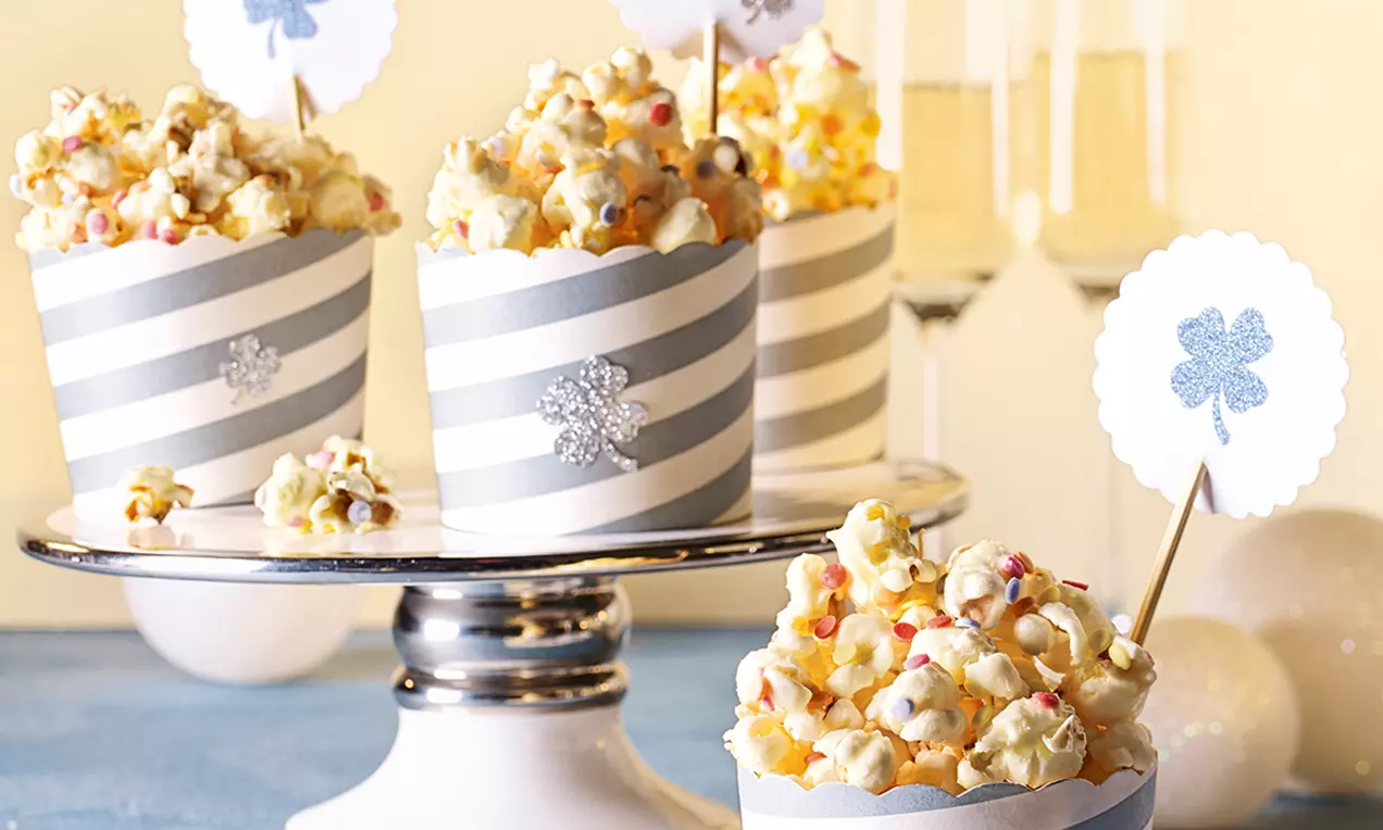 Weißes Popcorn mit glatter Schokoüberzug-Oberfläche und bunten Streuseln in gestreiften Papierbechern auf einer silbernen Etagere drapiert