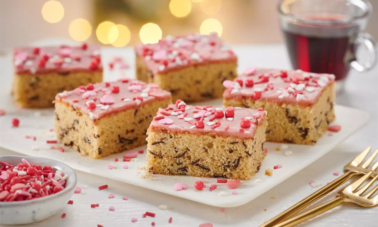 Glühweinkuchenwürfel mit rosafarbenem Guss und roten Zuckerstreuseln auf weißer Platte, daneben goldfarbene Gabeln und ein Glas Glühwein