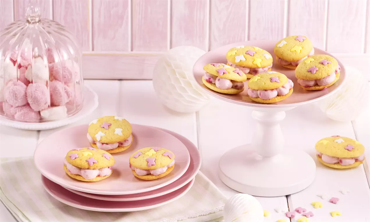 Goldgelbe, runde Whoopies mit zartrosa Himbeercremefüllung und Sternstreuseln auf rosafarbenen Tellern und weißer Etagere drapiert