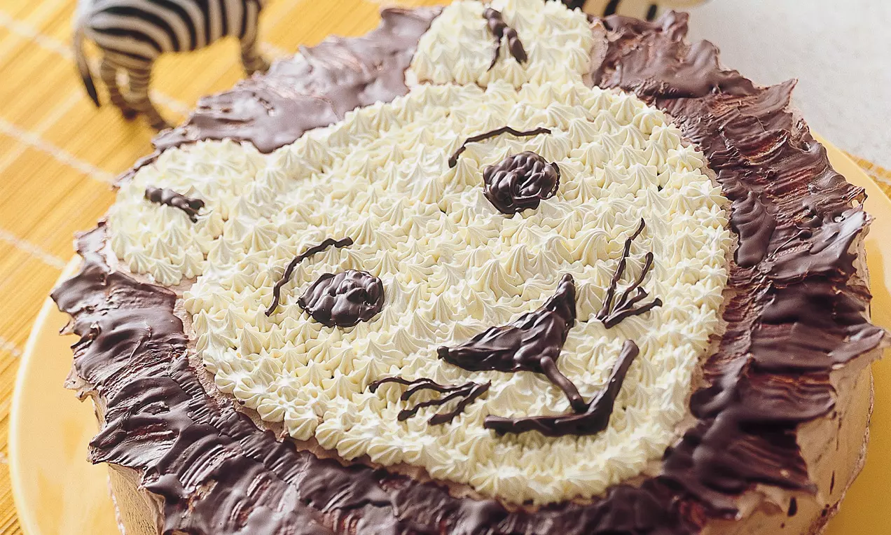 Runde Torte mit weißer Sahnecreme in Löwengesichtform, braunem Schokoladenhaar, auf gelbem Teller, Zebradekoration im Hintergrund