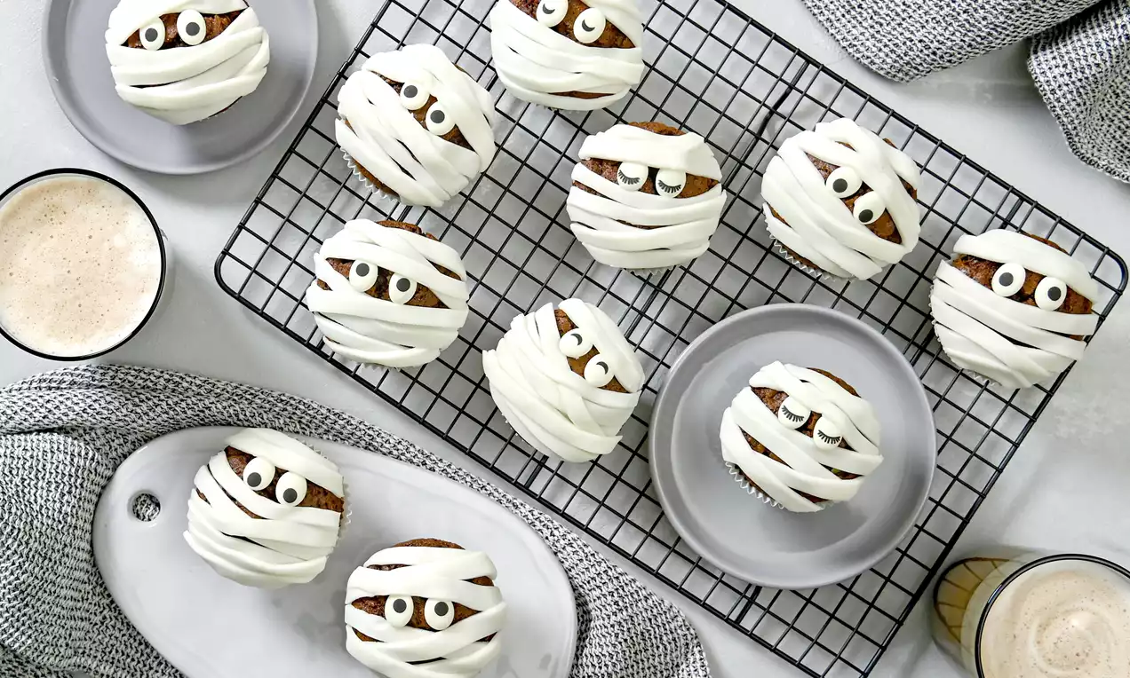 Hellbraune Muffins mit weißen Zuckerguss-Streifen als Mumien, dunklen Augen und weißer Glasur auf Gitter und grauen Tellern angerichtet