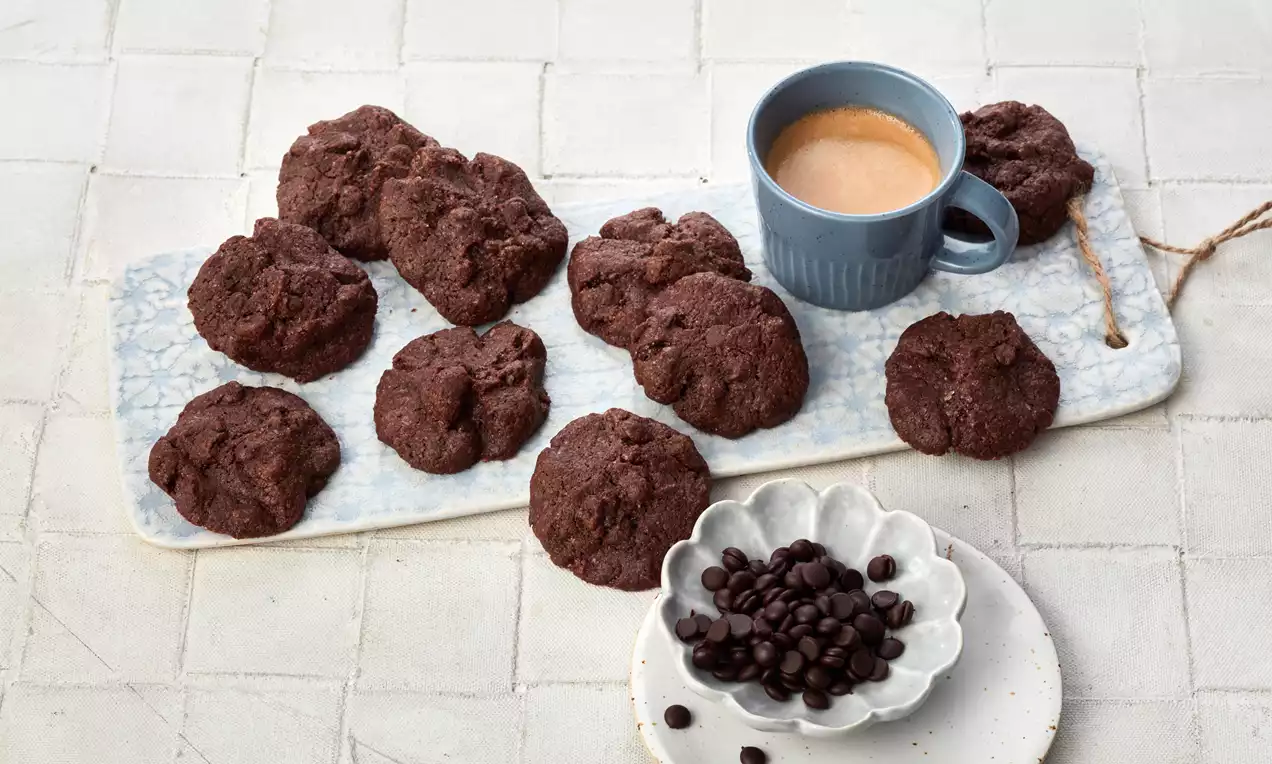 Dunkelbraune, grob strukturierte Schokoladen-Cookies auf länglicher Marmorplatte neben einer blauen Tasse Espresso und weißen Schalen mit Kaffeebohnen