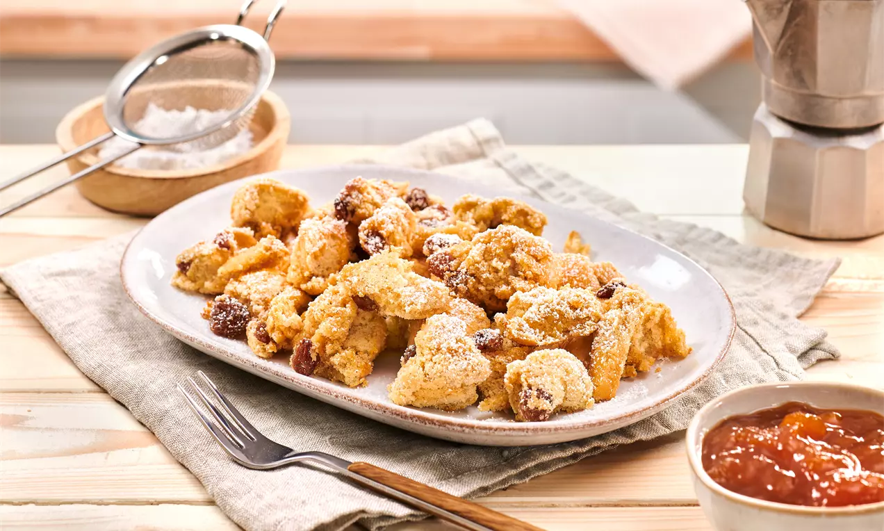 Goldbraune Kaiserschmarrn-Stücke mit Puderzucker bestäubt auf einem weißen Teller, daneben ein Schälchen Konfitüre und Besteck auf beigem Stoff