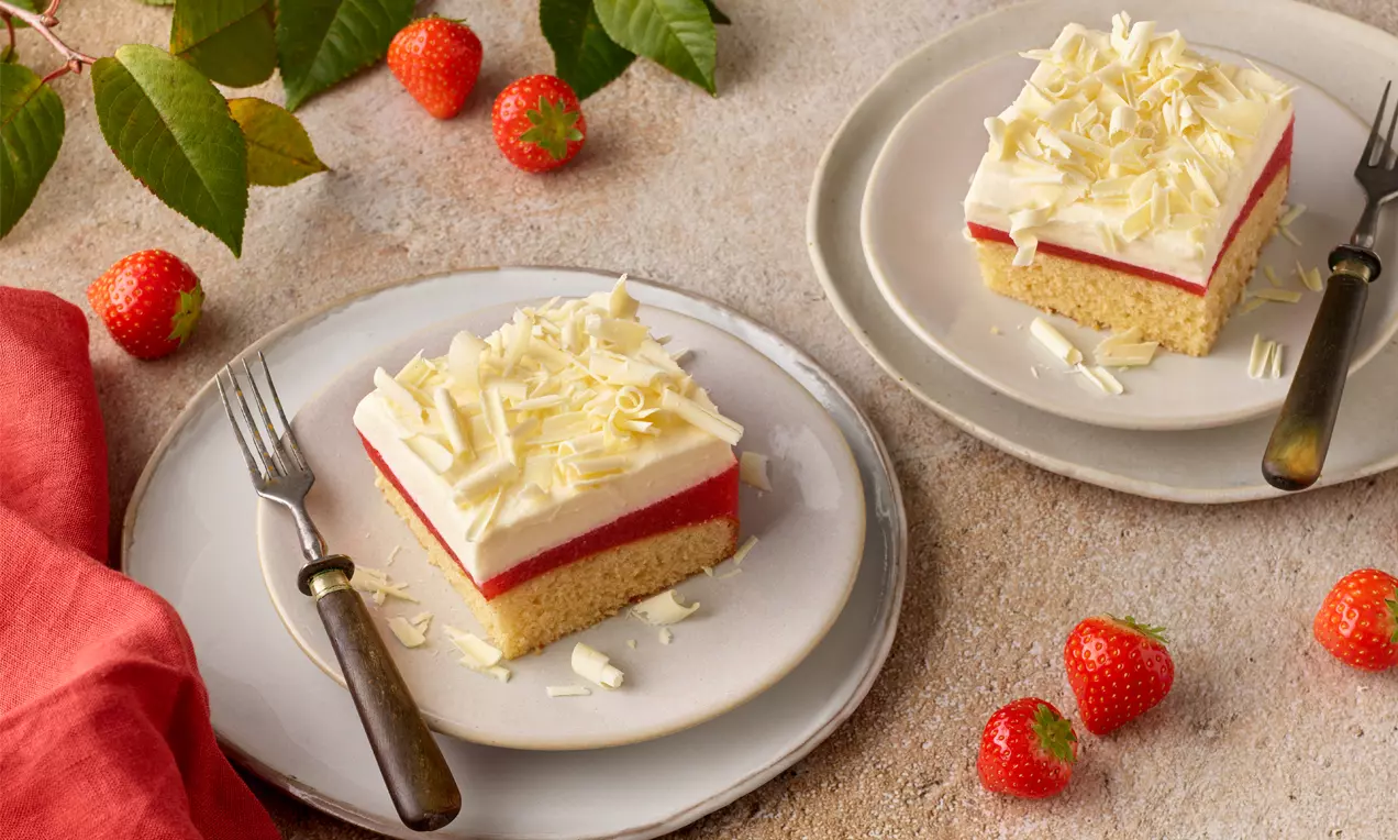Ein rechteckiges Stück Frau-Holle-Kuchen mit hellem Biskuitboden, roter Geleeschicht, weißer Cremeschicht und geraspelter weißer Schokolade
