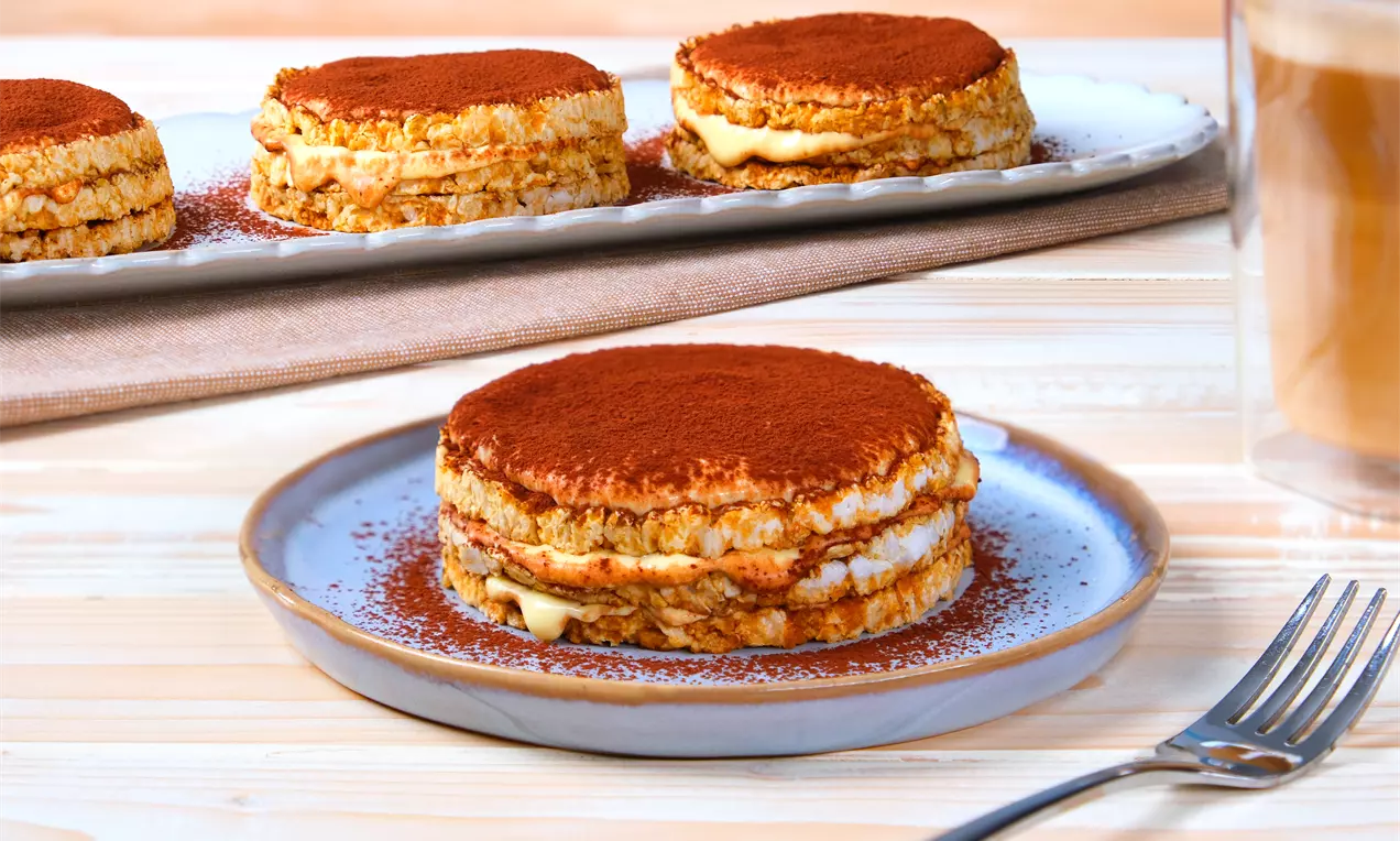 Reiswaffeltiramisu mit goldenem Knusperboden, heller Creme und Kakaopulver bestäubt auf hellblauem Teller, mit Gabel und Getränk